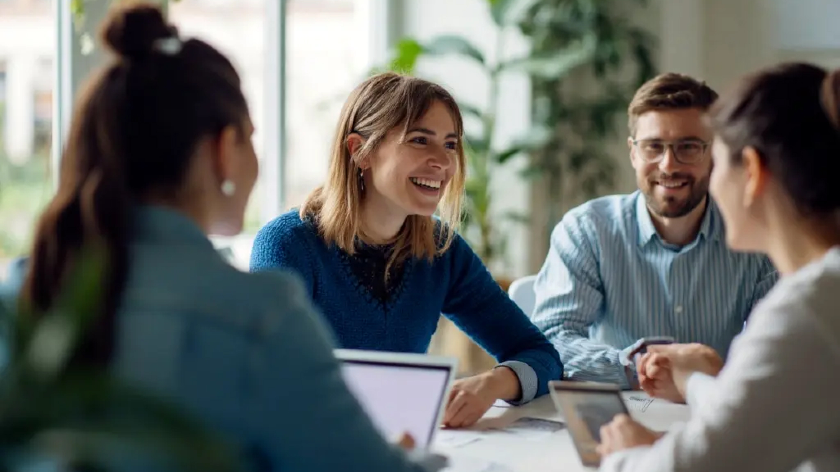 A group of colleagues sit together in a bright office, smiling and talking during a meeting.