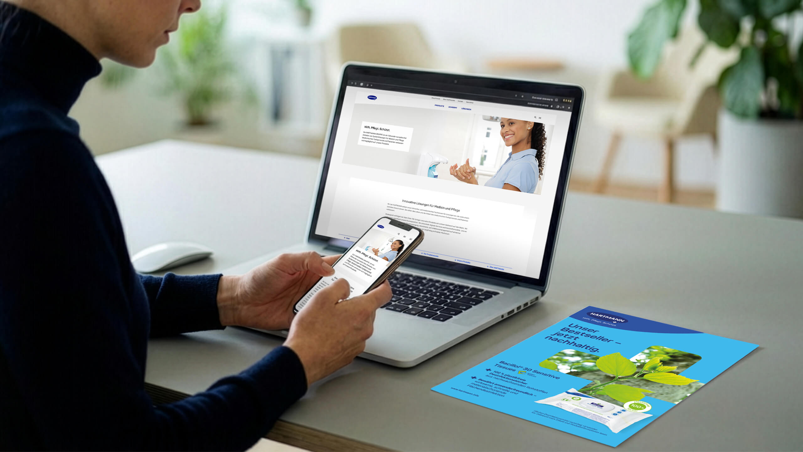 Person sitting at a desk using a smartphone while viewing the HARTMANN website on a laptop; a blue HARTMANN sustainability brochure lies on the table next to the computer.